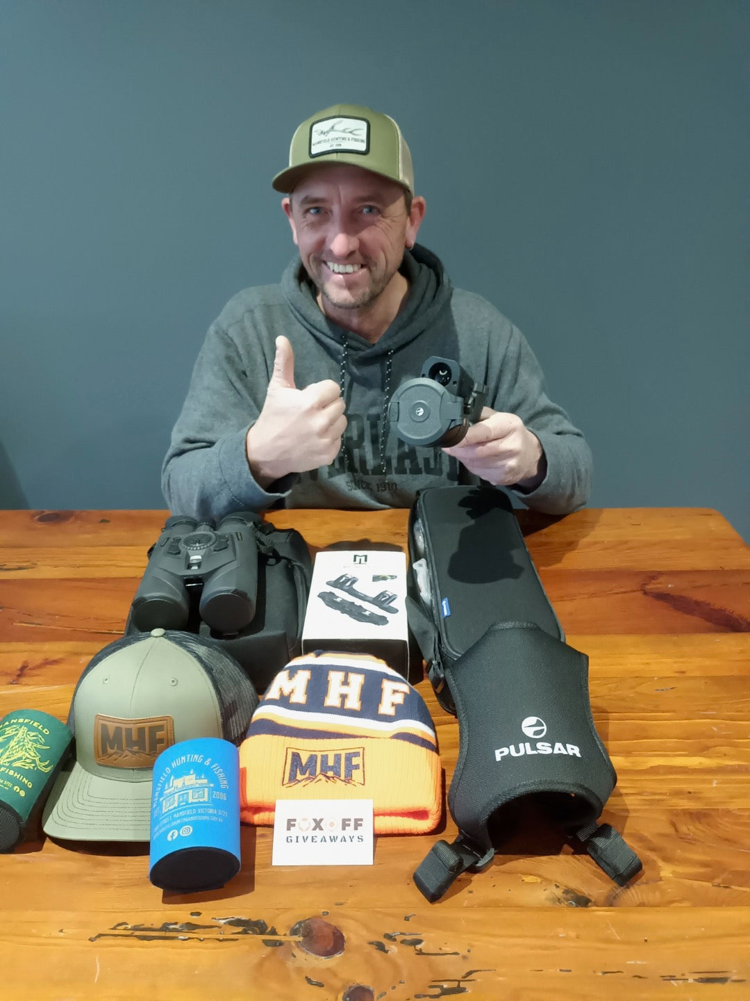 A man wearing a green cap and gray hoodie sits behind a wooden table displaying various outdoor gear, including a black Pulsar knee pad, a blue and yellow MUF fleece, a blue beanie, and other hats.
