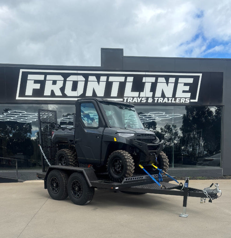 A black Polaris ATV sits on a black flatbed trailer parked outside a dealership building with a large 'FRONTLINE TRAYS & TRAILERS' sign. The trailer is hitched to a white pickup truck, and the scene is set on a concrete lot under a cloudy sky.
